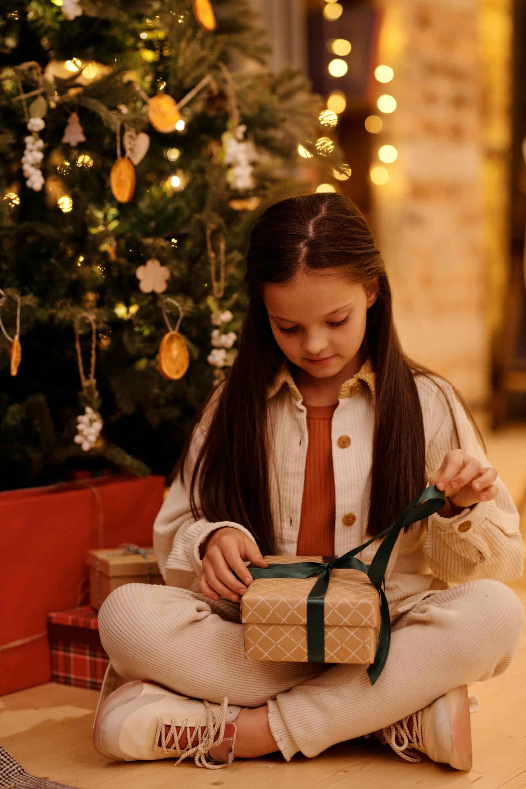 A young girl joyfully opens a Christmas present beside a decorated tree.